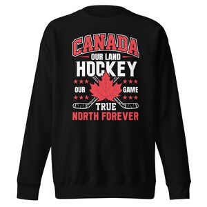 Pode incluir: Camisola de moletom preta com um design gráfico vermelho, branco e azul que diz "Canada Our Land Hockey Our True North Forever Game". O gráfico inclui uma folha de bordo e tacos de hóquei.