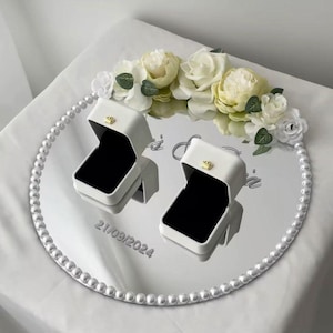 Puede incluir: Una bandeja redonda con espejo y borde de perlas sostiene dos joyeros blancos abiertos con interior negro. La bandeja está decorada con rosas blancas y crema y la fecha 21/09/2024.