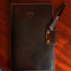 May include: A brown leather journal with a pen holder and a small leather flap with two brass rivets. The journal has a stitched edge and the text "Leather Goods" is embossed on the front cover.