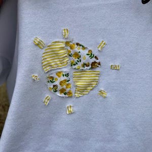May include: White hoodie with a yellow and white patchwork sun design. The sun is made of fabric scraps sewn together in a circular shape.