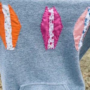 May include: A gray hoodie with a large pocket on the front. The hoodie has three diamond-shaped patches sewn on the front, each made of different patterned fabrics in pink, orange, and blue.