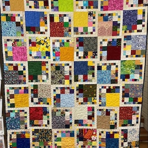 Può includere: Una trapunta colorata con un design a patchwork. Ogni quadrato è composto da quadrati più piccoli in vari colori e motivi. La trapunta è incorniciata da un bordo bianco e un bordo giallo.