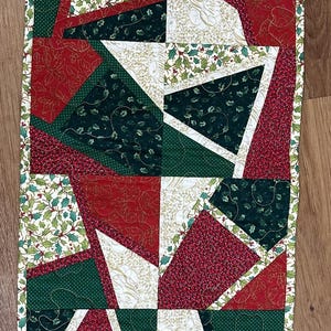 Puede incluir: Camino de mesa navideño rectangular con diseño de patchwork. El camino de mesa presenta telas rojas, verdes y blancas con motivos de acebo y geométricos. Los bordes están ribeteados con un estampado de acebo.