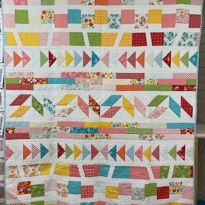May include: A colourful quilt with a white background and a variety of geometric patterns in shades of pink, red, yellow, green, blue, and turquoise. The quilt is made up of squares, rectangles, and triangles, and it has a modern, geometric design.