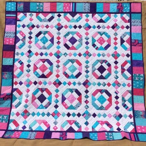 May include: A quilt with a white background and a pattern of pink, purple, and turquoise squares. The quilt is bordered with a strip of pink, purple, and turquoise fabric.