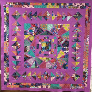May include: A colourful quilt with a purple background and a central square made up of smaller squares and triangles. The quilt is bordered by a row of triangles in various colours and patterns.