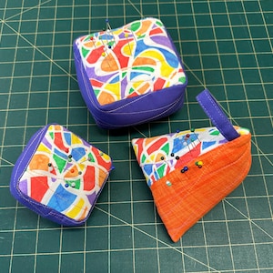 Könnte beinhalten: Drei Nadelkissen aus Stoff mit einem bunten abstrakten Muster aus roten, orangen, gelben, grünen und blauen Formen. Zwei sind quadratisch mit violetten Seiten, eines ist dreieckig mit einer orangen Seite und einer violetten Stoffschlaufe.