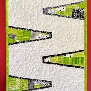 Può includere: Runner da tavolo trapuntato con sfondo bianco e forme geometriche verde lime e nere. Il runner è bordato con un tessuto verde lime. Le forme sono realizzate con vari tessuti fantasia.