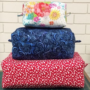 May include: Three fabric storage boxes stacked on top of each other. The top box is white with a floral print, the middle box is blue with a swirl pattern, and the bottom box is red with white polka dots.