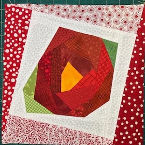 Könnte beinhalten: Ein quadratischer Quiltblock mit einem geometrischen Rosendesign. Die Rose besteht aus verschiedenen roten, orangefarbenen und grünen Stoffstücken. Der Block wird von weißem Stoff und rot gemusterten Rändern umrahmt. Der Text "Matilda's own" ist auf der rechten Seite sichtbar.
