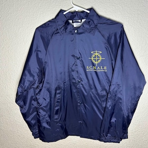 May include: Navy blue button-up jacket with a collar, hanging on a white hanger. The jacket has a yellow emblem on the left chest with the text "S.C.H.A.L.E. Independent Federal Investigation Club". The jacket is made of a shiny, lightweight material.