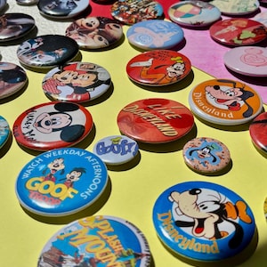May include: A collection of colorful vintage Disney character pinback buttons. The buttons feature images of Mickey Mouse, Goofy, and other characters, with text like "Disneyland" and "Goof Troop". The buttons are scattered on a yellow and red surface.