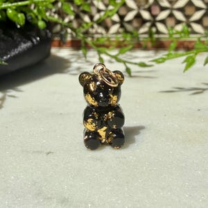 May include: A small, glossy black bear-shaped pendant featuring embedded gold-coloured flakes throughout its surface. The bear figure has rounded ears and a smooth, reflective finish, with a small gold-toned metal jump ring attached to the top.