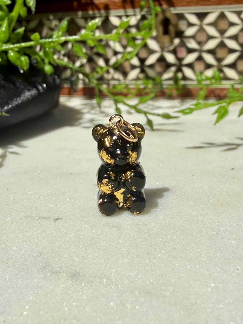 May include: A small, glossy black bear-shaped pendant featuring embedded gold-coloured flakes throughout its surface. The bear figure has rounded ears and a smooth, reflective finish, with a small gold-toned metal jump ring attached to the top.