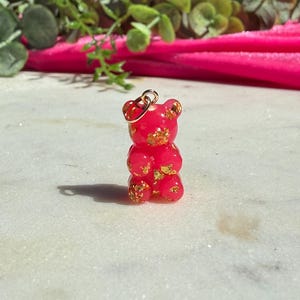May include: A small, translucent hot pink gummy bear pendant featuring embedded gold-coloured flakes throughout the body. The bear has a rounded head, small ears, and a smooth surface, with a gold-toned metal jump ring attached to the top of its head.