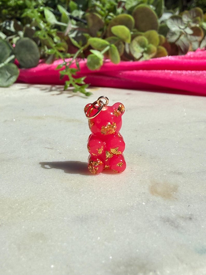 May include: A small, translucent hot pink gummy bear pendant featuring embedded gold-coloured flakes throughout the body. The bear has a rounded head, small ears, and a smooth surface, with a gold-toned metal jump ring attached to the top of its head.