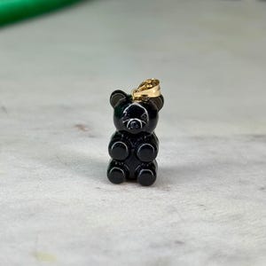 May include: A small, black gummy bear pendant with a gold-coloured bail. The bear is glossy and has a classic gummy bear shape. The pendant is likely made of a resin or similar material. The background is a light grey surface.