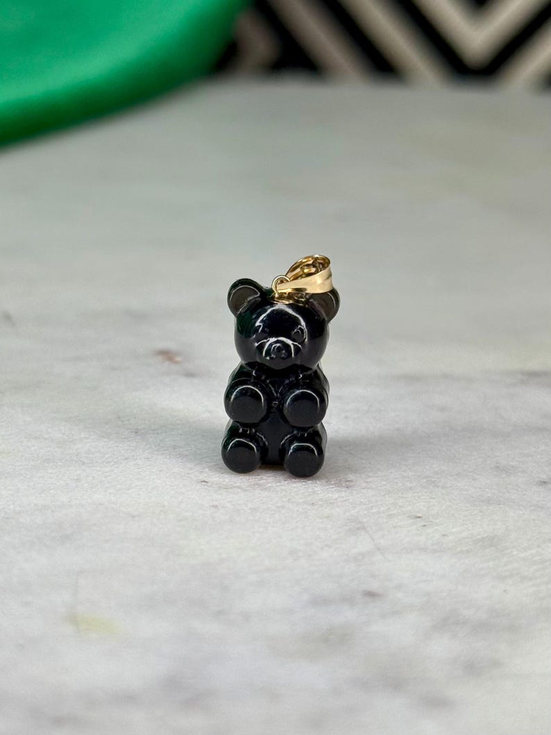 May include: A small, black gummy bear pendant with a gold-coloured bail. The bear is glossy and has a classic gummy bear shape. The pendant is likely made of a resin or similar material. The background is a light grey surface.