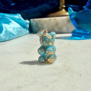 May include: A small, light blue resin bear pendant shaped like a gummy candy, featuring embedded gold leaf flakes throughout the translucent material. A simple gold-toned metal jump ring is attached to the top of the bear's head for hanging.