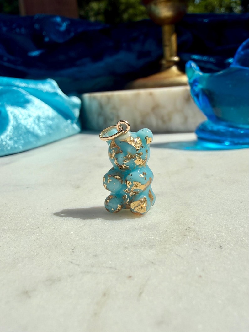May include: A small, light blue resin bear pendant shaped like a gummy candy, featuring embedded gold leaf flakes throughout the translucent material. A simple gold-toned metal jump ring is attached to the top of the bear's head for hanging.