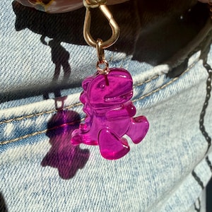 May include: A translucent, vibrant magenta dinosaur-shaped charm made of resin, suspended from a gold-toned metal lobster clasp. The dinosaur figure features a smooth, glossy finish and a rounded, cartoon-like design with a visible tail and limbs.
