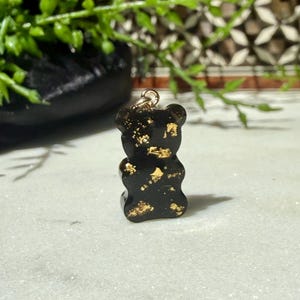 May include: A small, black, bear-shaped pendant made of resin, featuring embedded gold-coloured metallic flakes throughout its surface. The bear figure has a smooth, glossy finish and includes a small, circular gold-toned metal jump ring attached to the top for hanging.