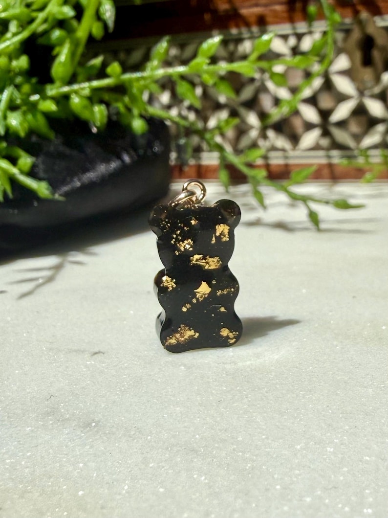 May include: A small, black, bear-shaped pendant made of resin, featuring embedded gold-coloured metallic flakes throughout its surface. The bear figure has a smooth, glossy finish and includes a small, circular gold-toned metal jump ring attached to the top for hanging.