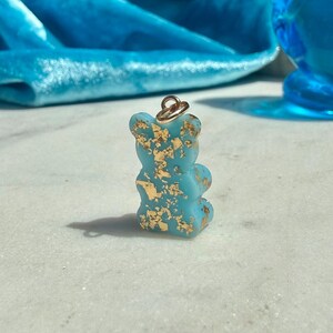 May include: A small, opaque light blue bear-shaped pendant featuring embedded gold-coloured metallic flakes throughout its surface. The bear figure includes a small gold-toned metal jump ring attached to the top for hanging as a jewellery charm.