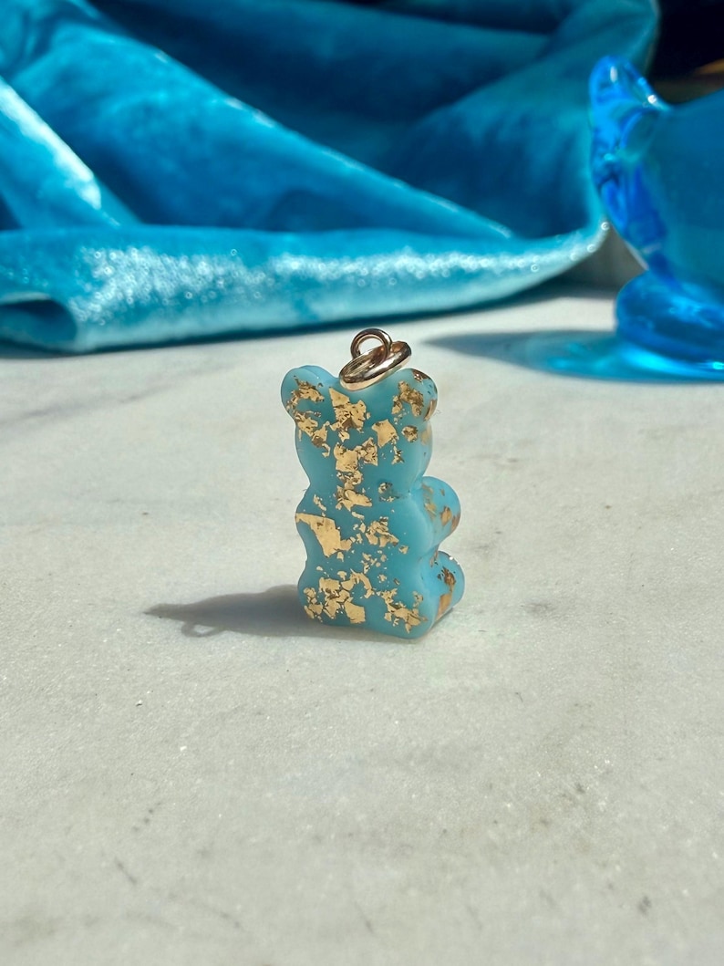 May include: A small, opaque light blue bear-shaped pendant featuring embedded gold-coloured metallic flakes throughout its surface. The bear figure includes a small gold-toned metal jump ring attached to the top for hanging as a jewellery charm.