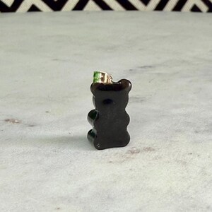 May include: A small, dark brown gummy bear pendant with a gold-coloured bail and a green accent on top. The pendant is set against a white and grey marble surface with a black and white geometric pattern in the background.