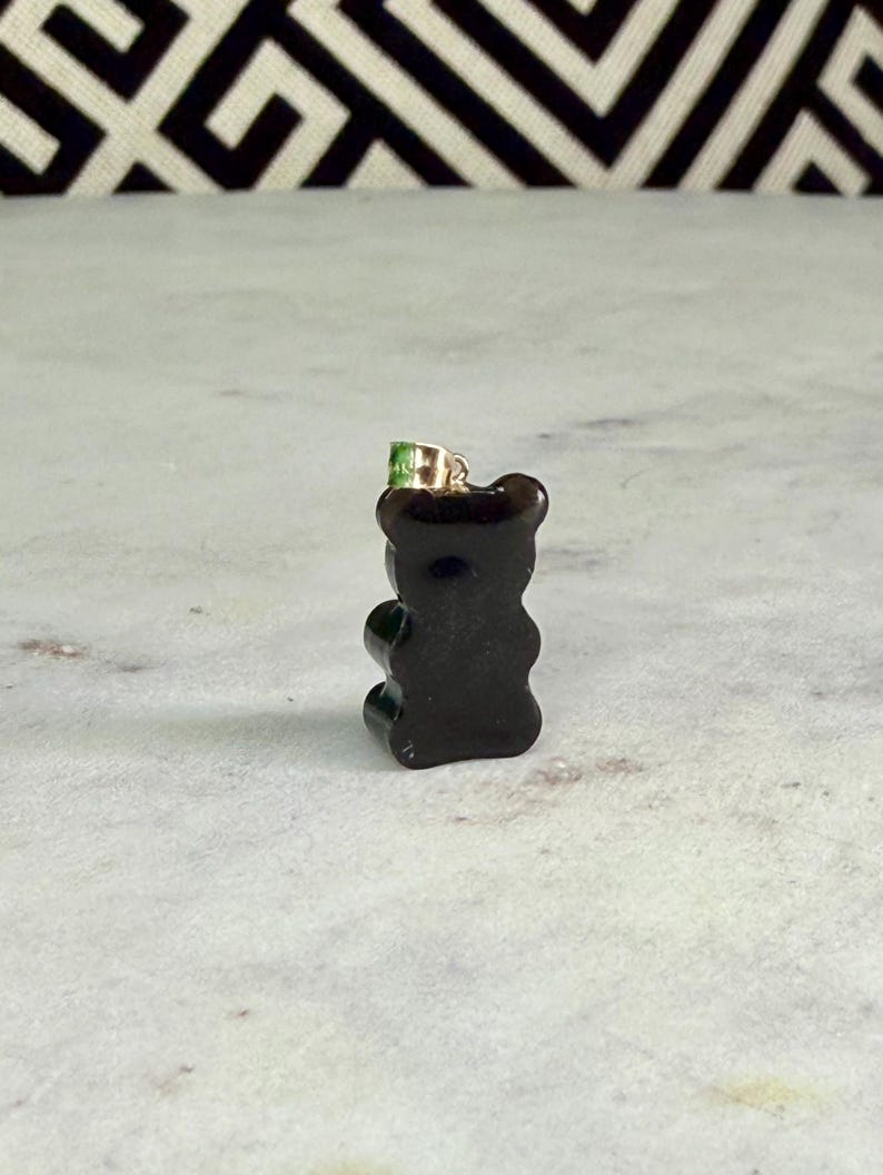 May include: A small, dark brown gummy bear pendant with a gold-coloured bail and a green accent on top. The pendant is set against a white and grey marble surface with a black and white geometric pattern in the background.