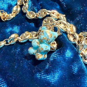 May include: A gold-toned chain necklace featuring a small, light blue gummy bear pendant with embedded gold-coloured flakes. The pendant is attached to the chain with a small gold-toned jump ring, creating a contrast between the metallic chain and the bear.