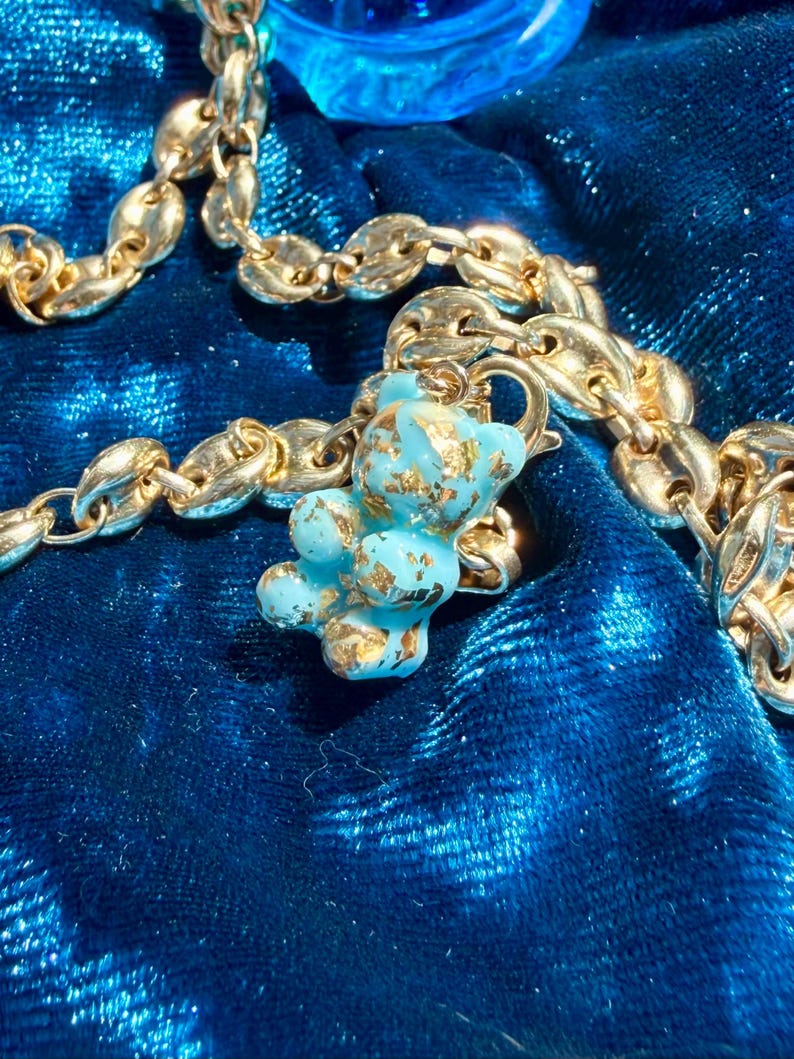 May include: A gold-toned chain necklace featuring a small, light blue gummy bear pendant with embedded gold-coloured flakes. The pendant is attached to the chain with a small gold-toned jump ring, creating a contrast between the metallic chain and the bear.