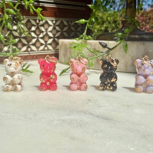 May include: Five small gummy bear charms arranged in a row, each featuring a different solid colour—white, hot pink, light pink, black, and lavender—with embedded gold foil flakes and a small gold-toned metal jump ring attached to the top of each.