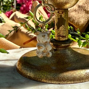 May include: A small, translucent white bear-shaped pendant featuring embedded gold-coloured flakes. The bear charm is attached to a gold-toned metal jump ring, which is connected to a gold-toned chain link, suspended from a decorative gold stand.