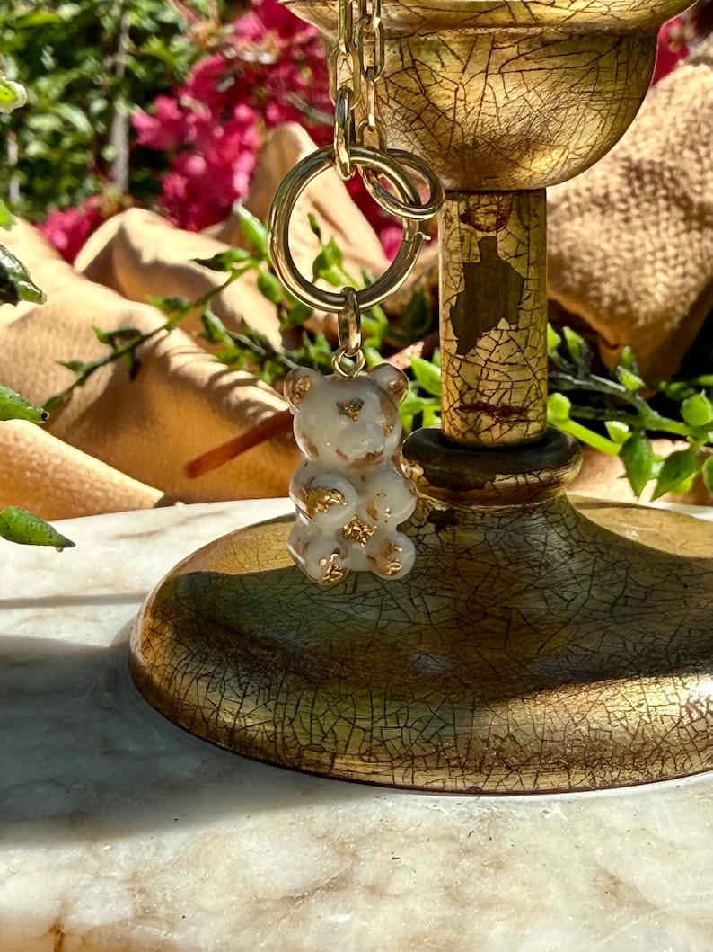 May include: A small, translucent white bear-shaped pendant featuring embedded gold-coloured flakes. The bear charm is attached to a gold-toned metal jump ring, which is connected to a gold-toned chain link, suspended from a decorative gold stand.