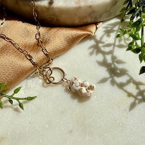 May include: A gold-toned chain necklace featuring a small, white, bear-shaped pendant with gold-coloured speckles. The pendant is attached to a circular gold-toned ring connector that links to the chain, creating a layered, delicate jewellery piece.