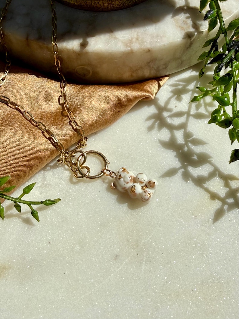 May include: A gold-toned chain necklace featuring a small, white, bear-shaped pendant with gold-coloured speckles. The pendant is attached to a circular gold-toned ring connector that links to the chain, creating a layered, delicate jewellery piece.