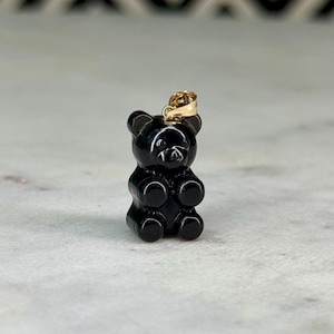 May include: A black gummy bear pendant with a gold-coloured bail. The pendant is a miniature, three-dimensional representation of a gummy bear. The bear has a smooth, glossy finish and is standing upright.