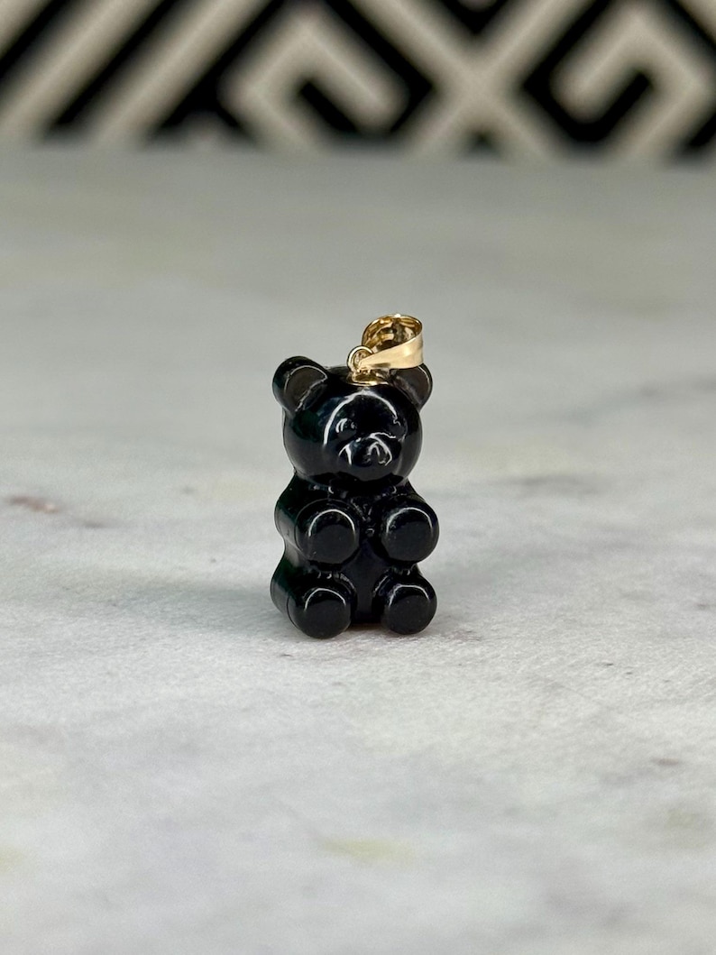 May include: A black gummy bear pendant with a gold-coloured bail. The pendant is a miniature, three-dimensional representation of a gummy bear. The bear has a smooth, glossy finish and is standing upright.