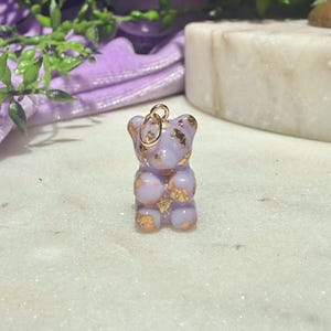 May include: A small, light purple, translucent bear-shaped pendant featuring embedded gold-coloured metallic flakes throughout its body. The bear figure has a smooth, glossy finish and includes a small, circular gold-toned metal jump ring at the top.