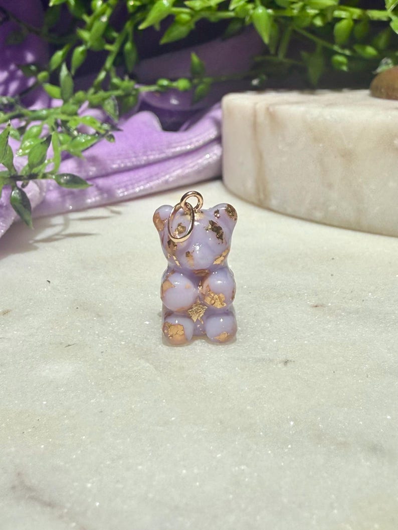 May include: A small, light purple, translucent bear-shaped pendant featuring embedded gold-coloured metallic flakes throughout its body. The bear figure has a smooth, glossy finish and includes a small, circular gold-toned metal jump ring at the top.