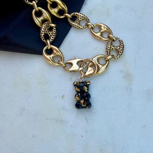 May include: A gold-toned chain necklace featuring alternating smooth and textured oval links. A small, black bear-shaped pendant with gold-coloured flecks is attached to the chain by a circular clasp, creating a distinct contrast between the metal links.
