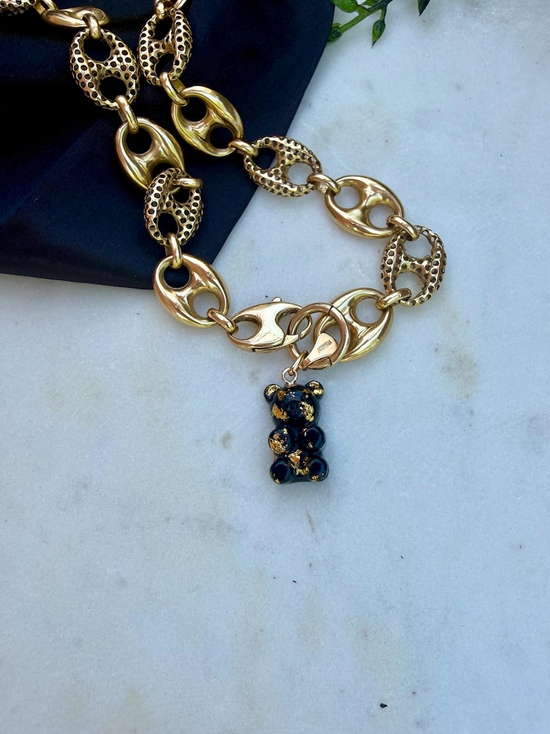 May include: A gold-toned chain necklace featuring alternating smooth and textured oval links. A small, black bear-shaped pendant with gold-coloured flecks is attached to the chain by a circular clasp, creating a distinct contrast between the metal links.