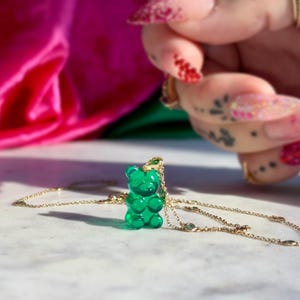 May include: A gold necklace featuring a green gummy bear pendant. The pendant is a bright, glossy green. The necklace rests on a white marble surface, with a pink and green backdrop. The necklace is a piece of jewellery.