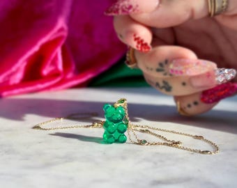 May include: A gold-colored necklace with a green gummy bear pendant. The gummy bear is a vibrant green color and has a glossy finish. The necklace is on a white marble surface, with a pink and green background.