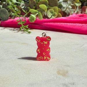 May include: A small, translucent red gummy bear pendant featuring embedded gold-coloured metallic flakes throughout the resin material. The bear-shaped charm includes a small silver-toned metal jump ring attached to the top for hanging purposes.