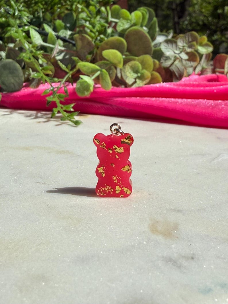 May include: A small, translucent red gummy bear pendant featuring embedded gold-coloured metallic flakes throughout the resin material. The bear-shaped charm includes a small silver-toned metal jump ring attached to the top for hanging purposes.