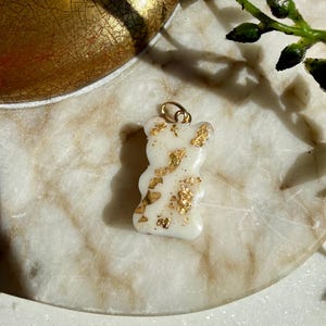 May include: A small, opaque white bear-shaped pendant featuring embedded gold-coloured metallic flakes throughout its surface. The charm includes a small gold-toned metal jump ring attached to the top, designed for use as a jewellery component.