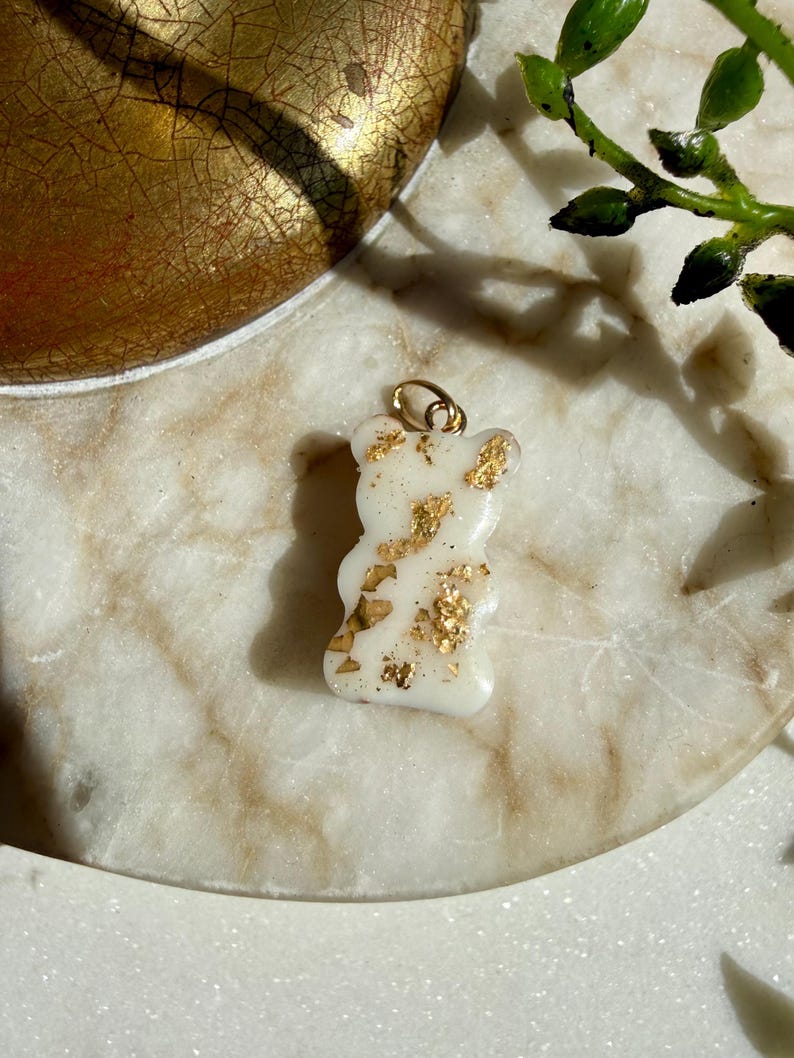 May include: A small, opaque white bear-shaped pendant featuring embedded gold-coloured metallic flakes throughout its surface. The charm includes a small gold-toned metal jump ring attached to the top, designed for use as a jewellery component.